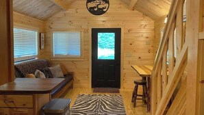 A cozy cabin interior with a wooden ceiling, a black door, a sofa, a dining table, and a zebra-patterned rug. - Asheboro 1BR Cabin Near NC Zoo | Loft & Water View (Asheboro)