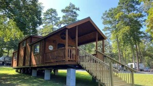 A wooden cabin with a balcony and a staircase leading up to it, situated on a grassy area with trees in the background. - Asheboro 1BR Cabin Near NC Zoo | Loft & Water View (Asheboro)