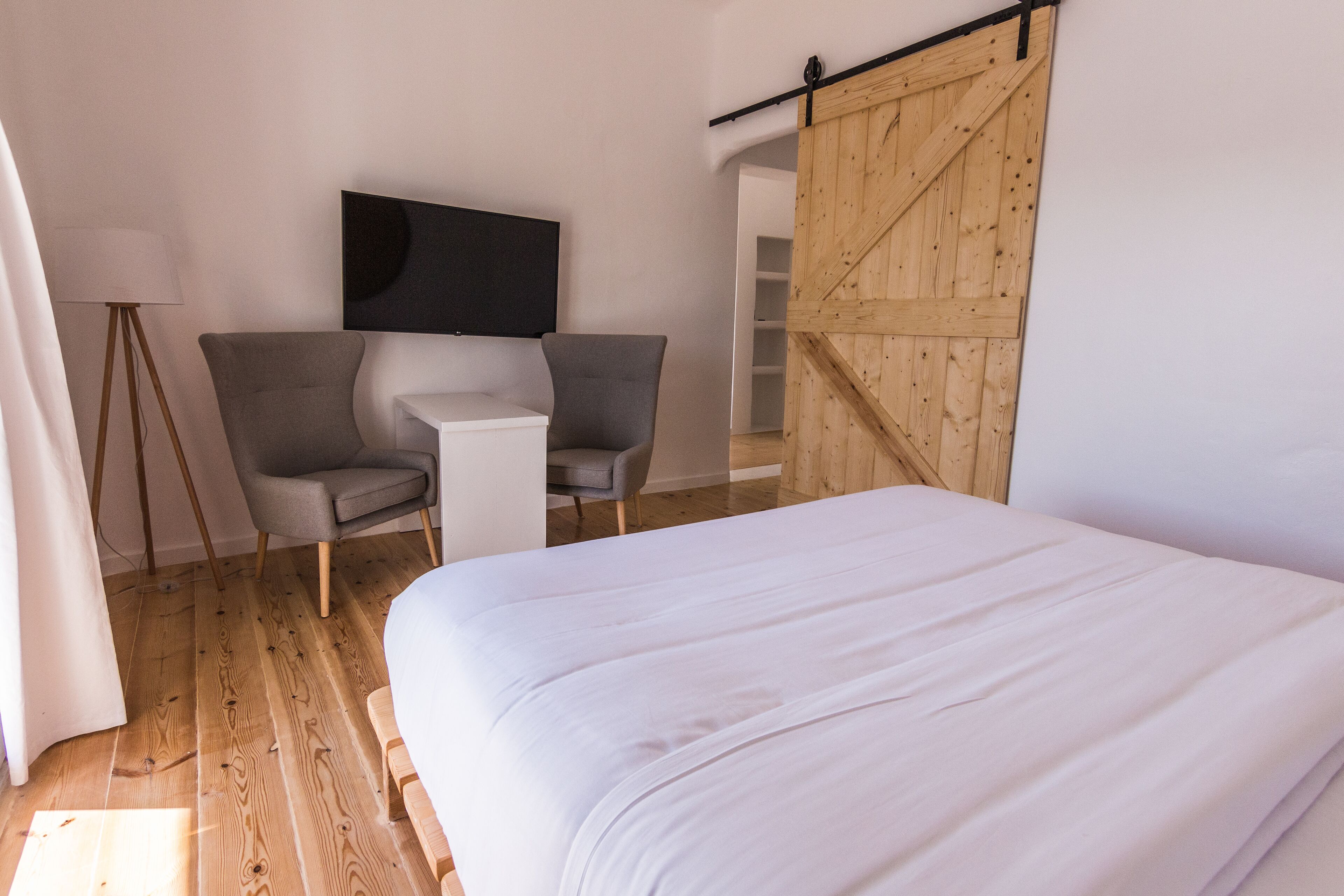 A bedroom with a large bed, a desk, two chairs, a TV mounted on the wall, and a wooden sliding door.