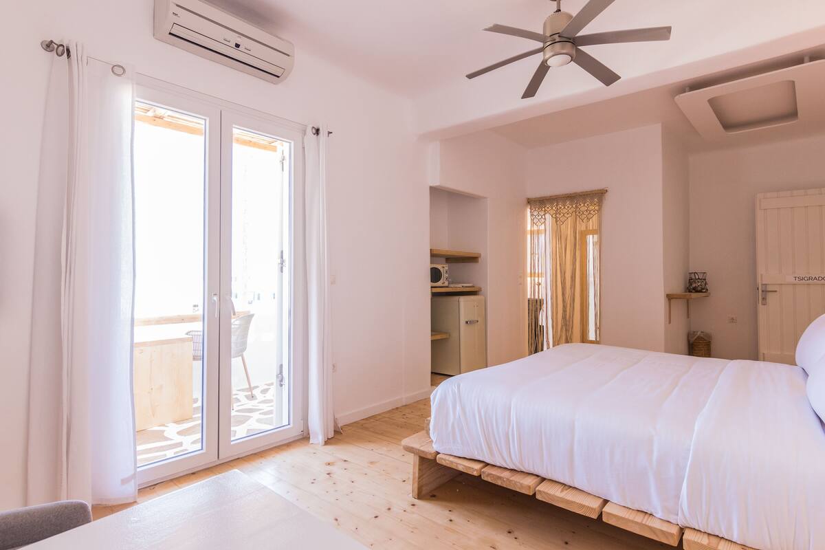 A bedroom with a bed, a ceiling fan, a sliding glass door, and a small kitchenette.