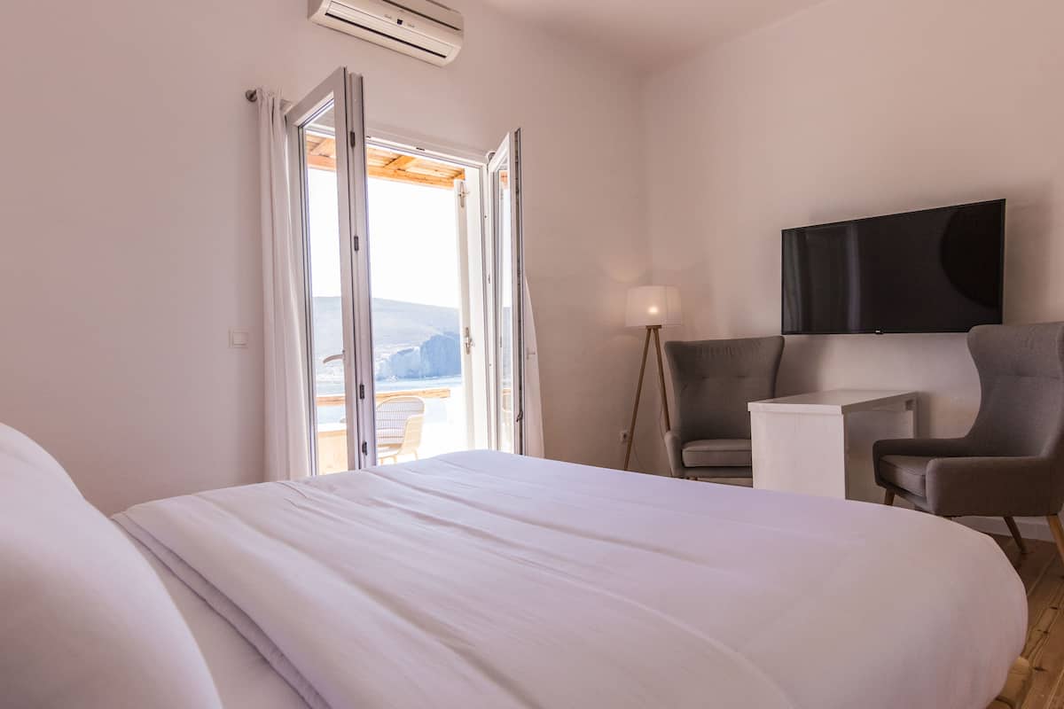 A hotel room with a bed, a chair, a desk, a TV, and a view of the sea through an open door.