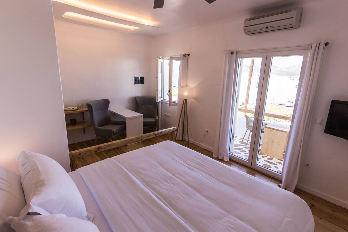 A hotel room with a large bed, a desk, a chair, and a view through the glass door.