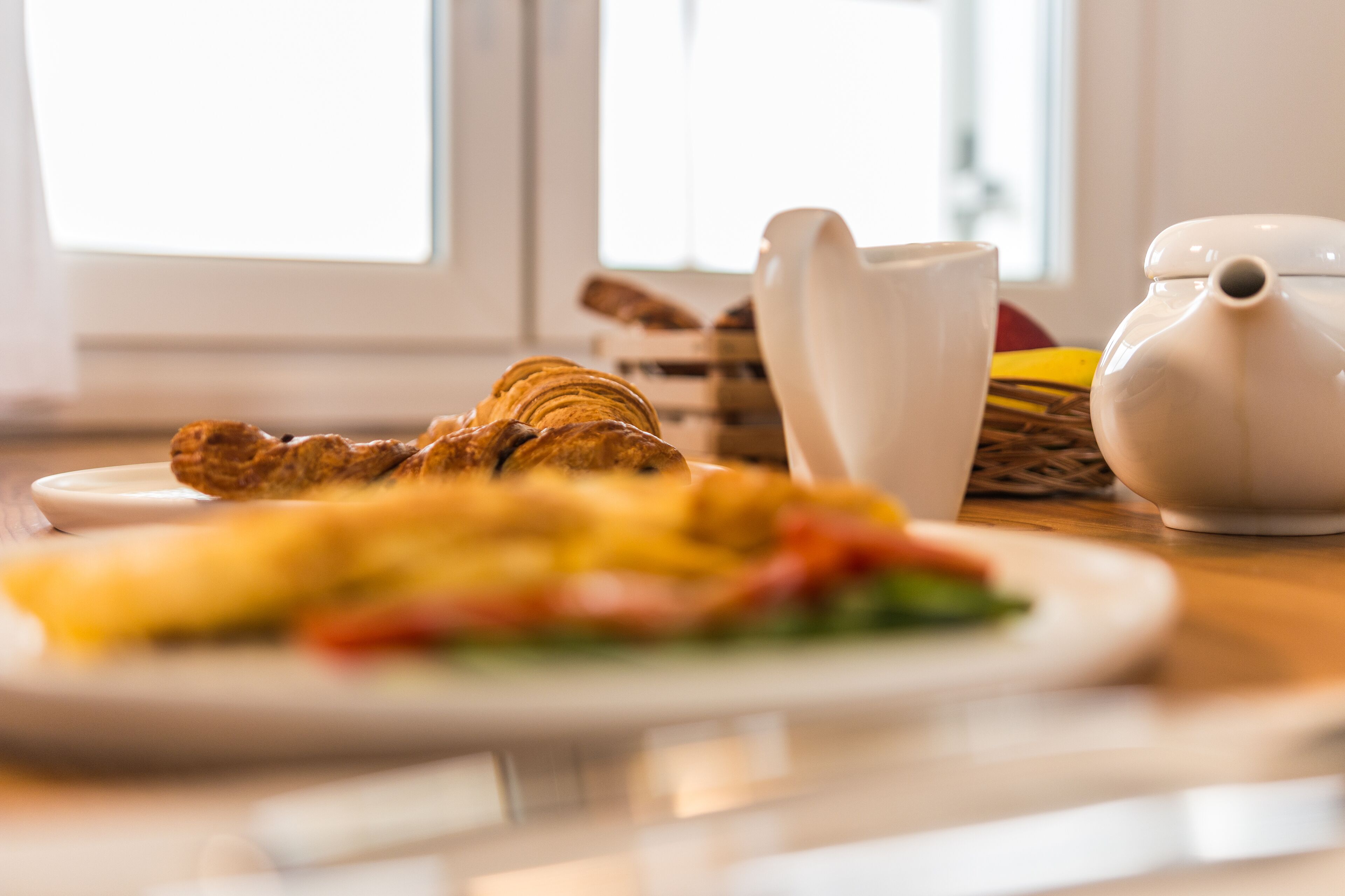 Una escena de desayuno con croissants, un plato de comida, una jarra blanca y una tetera sobre una mesa de madera cerca de una ventana.