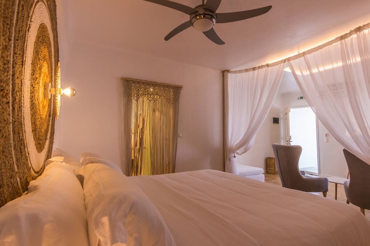 A bedroom with a large bed, a ceiling fan, sheer curtains, and a view of a balcony.