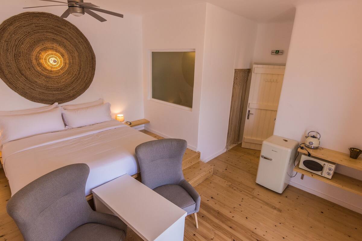 A bedroom with a bed, two chairs, a small table, a ceiling fan, a wall-mounted light, a window, a microwave, and a wooden floor.