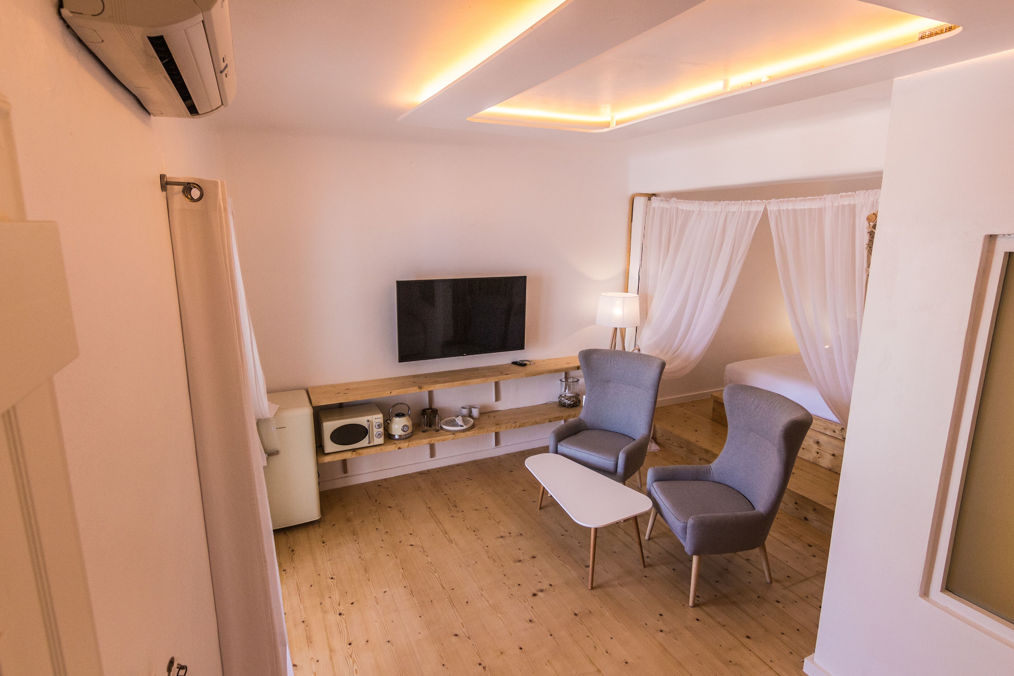 Una habitación de hotel moderna con un televisor de pantalla plana, una consola de madera con varios objetos, dos sillones grises, una mesita blanca pequeña y una cama con dosel.