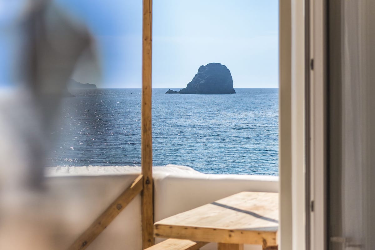 An open door leading to a balcony with a wooden table and benches, overlooking a calm sea and a distant island.