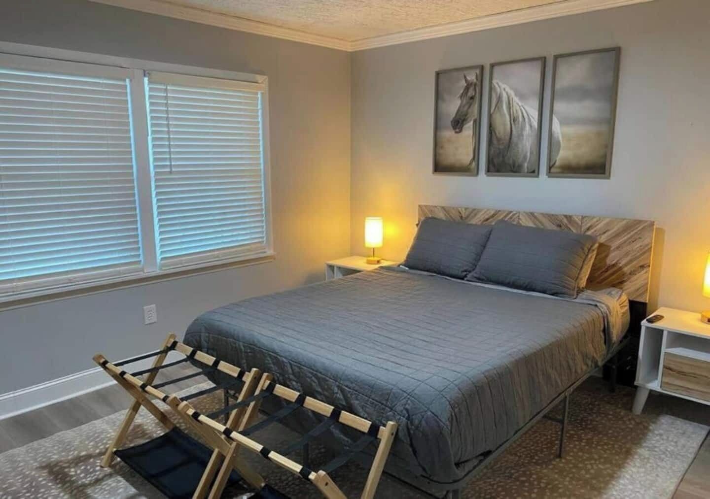A bedroom with a bed, two bedside tables, a window with blinds, and three horse paintings on the wall.