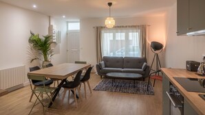 Una sala moderna con una mesa de comedor de madera, sillas negras, un sofá gris y una pequeña mesa de centro. Hay una planta en maceta, una lámpara de pie y una cocina con estufa y armarios.