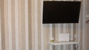 A room with a striped wallpaper, a mounted TV, a desk with a computer, and a patterned bedspread. - Renovated Studio Olympiados (Thessaloniki)