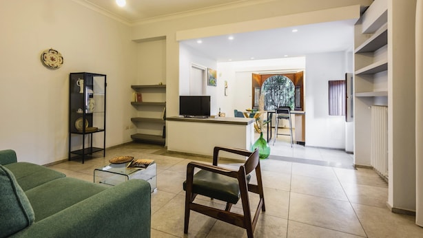 A modern living room with a green sofa, a wooden chair, and a glass coffee table. There is a kitchen area in the background with a TV and a dining table. - Euphorbia Apartment 2 in Catania by Wonderful Italy (Catania)