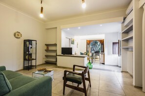 A modern living room with a green sofa, a wooden chair, and a glass coffee table. There is a kitchen area in the background with a TV and a dining table. - Euphorbia Apartment 2 in Catania by Wonderful Italy (Catania)