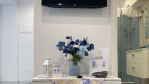 Une chambre d’hôtel avec un téléviseur à écran plat fixé au mur, une petite table sur laquelle est posé un vase de fleurs, une enseigne indiquant « La Piccola Suite », et un sol carrelé.