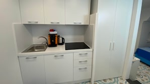 Une petite cuisine équipée comprenant un évier, une machine à café et une bouilloire. On y trouve des placards blancs avec des tiroirs, ainsi qu’un grand placard blanc sur le côté droit.