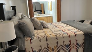 Ein Schlafzimmer mit Bett, Nachttisch, Lampe und Blick auf das Badezimmer sowie die Küche durch einen Bogen.