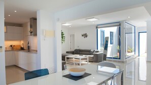Una cocina moderna con armarios blancos, horno empotrado y cafetera. Hay un comedor con mesa y sillas de vidrio, y una sala de estar con sofá gris y una mesa de centro.