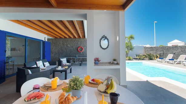 Zona de comedor junto a la piscina, con una mesa preparada para el desayuno, incluyendo cruasanes, fruta y zumo.