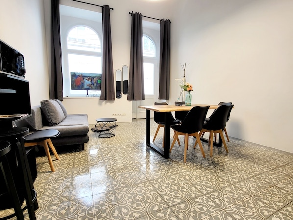 Ein modernes Wohnzimmer mit einem grauen Sofa, einem kleinen schwarzen Tisch und einem Essbereich mit einem Holztisch und schwarzen Stühlen.
