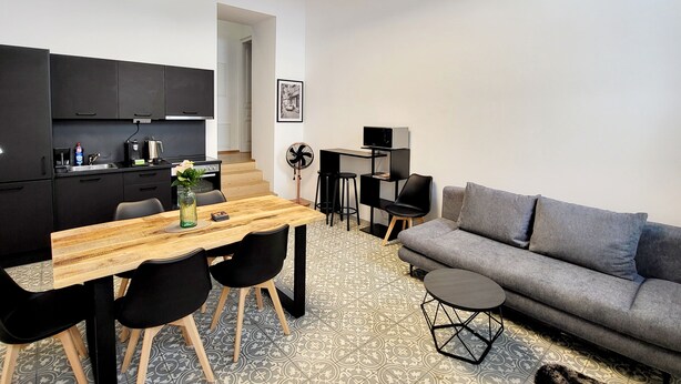 Eine moderne Küche mit schwarzen Schränken, einem Holztisch und schwarzen Stühlen. Im Wohnbereich befindet sich eine graue Couch und ein kleiner runder Tisch.