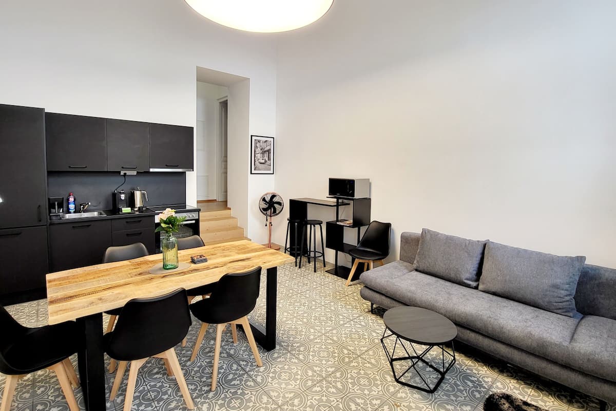 A modern kitchen with black cabinets, a wooden dining table, and black chairs. There is a grey sofa and a small round table in the living area.