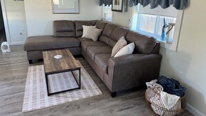 A modern living room with a sectional sofa, a coffee table, and a large window with curtains. - Quaint 1 Bed 1 bath private beach home. Long term renter welcome Dec 1 (Dartmouth)