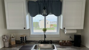 A kitchen with white cabinets, a stainless steel sink, a coffee machine, and a window with a view of a house and greenery. - Quaint 1 Bed 1 bath private beach home. Long term renter welcome Dec 1 (Dartmouth)