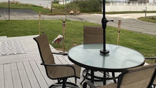 A deck with a glass-top table and four chairs, an umbrella, and a view of a road and buildings in the distance. - Quaint 1 Bed 1 bath private beach home. Long term renter welcome Dec 1 (Dartmouth)