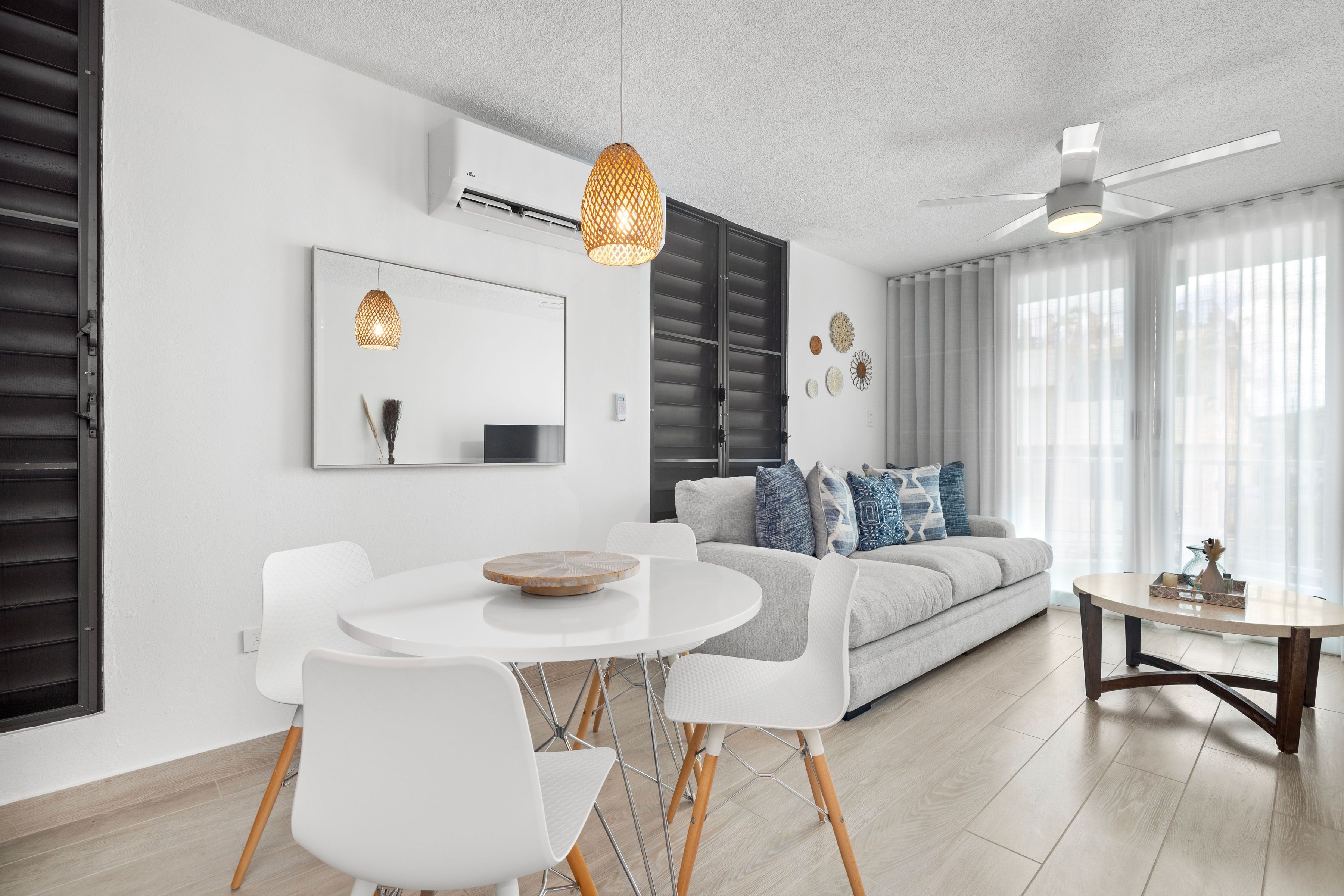 Una sala moderna con un sofá blanco, una mesa de comedor con sillas, un ventilador de techo y un ventanal con cortinas traslúcidas.