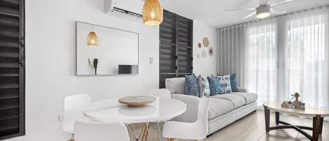 Una sala moderna con un sofá blanco, una mesa de comedor con sillas, un ventilador de techo y un ventanal con cortinas traslúcidas.