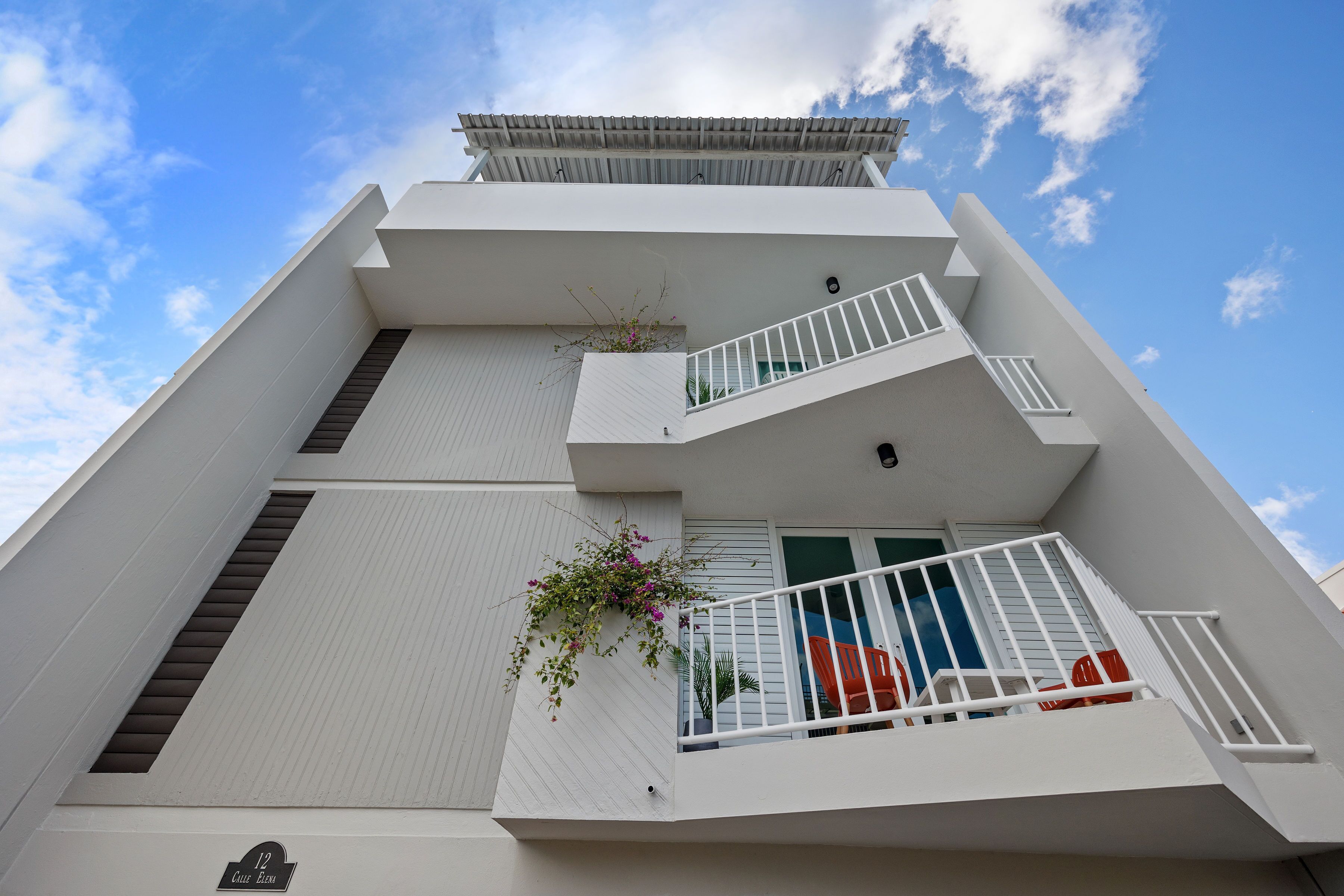 Un edificio moderno con fachada blanca, un balcón con barandilla y un pequeño jardín con plantas.