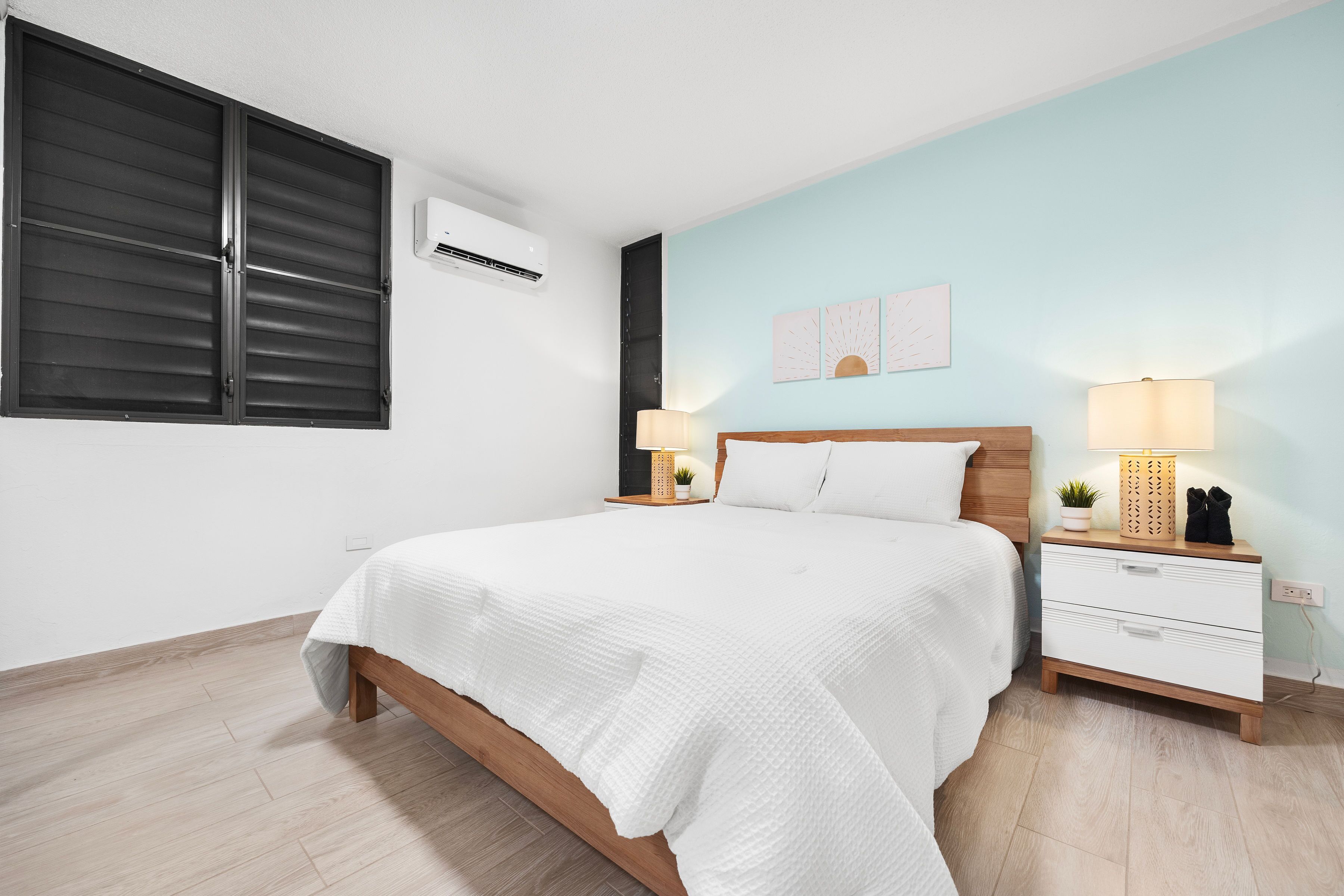 A bedroom with a bed, bedside tables, a nightstand, an air conditioning unit, and a wall with two decorative pieces.