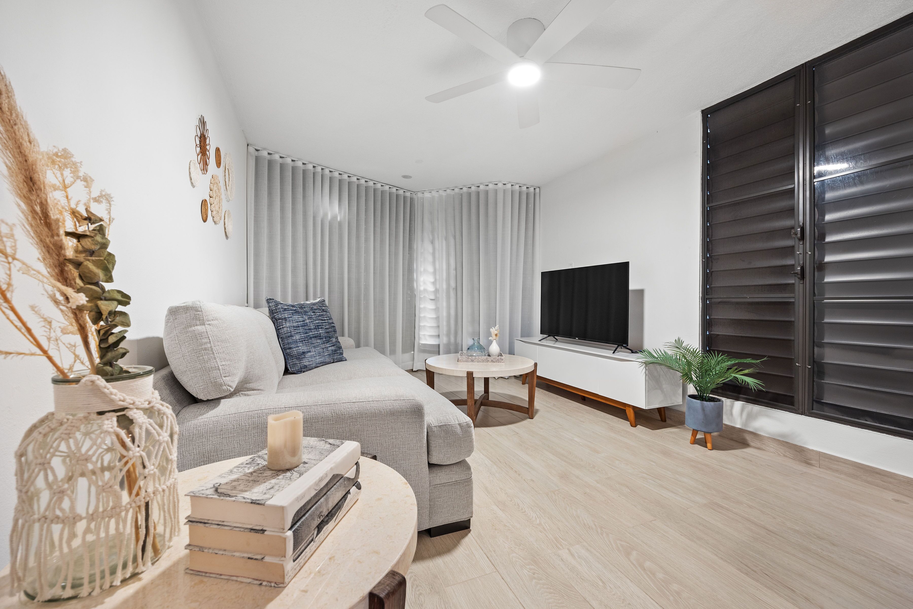 A modern living room with a grey sofa, a small round table, a TV, and a large window with blinds.
