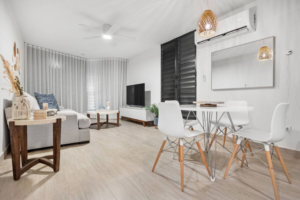 Una sala de estar moderna con sofá, mesa de centro y zona de comedor con sillas. Hay un televisor, una planta y un ventilador de techo.