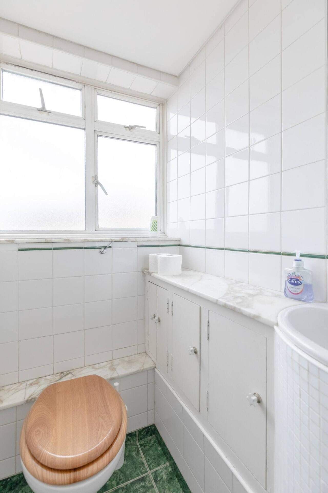 A small, white bathroom with a wooden toilet, a window, and a white sink with a soap dispenser.