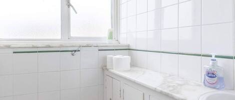 A small, white bathroom with a wooden toilet, a window, and a white sink with a soap dispenser.