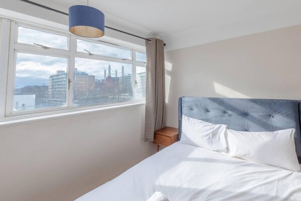 A bedroom with a large window, a bed with white linens, a blue headboard, and a wooden nightstand.