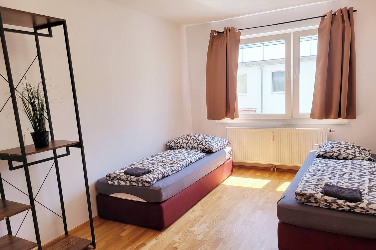 A compact room with two single beds, a wooden floor, a radiator, and a window with curtains.