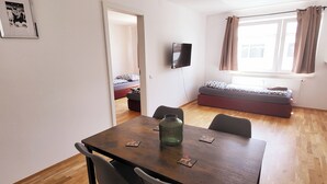 A compact living space with a dining area, a TV mounted on the wall, a bed with patterned bedding, and a window with curtains.