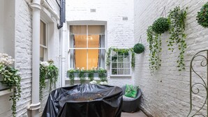 A narrow courtyard with a covered table, a window with curtains, and hanging plants. - Elegant 2BD Flat 5 Mins to Kensington Gardens! (London)