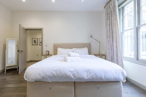 A neatly made bed with white linens, a bedside lamp, and a window with curtains. - Elegant 2BD Flat 5 Mins to Kensington Gardens! (London)