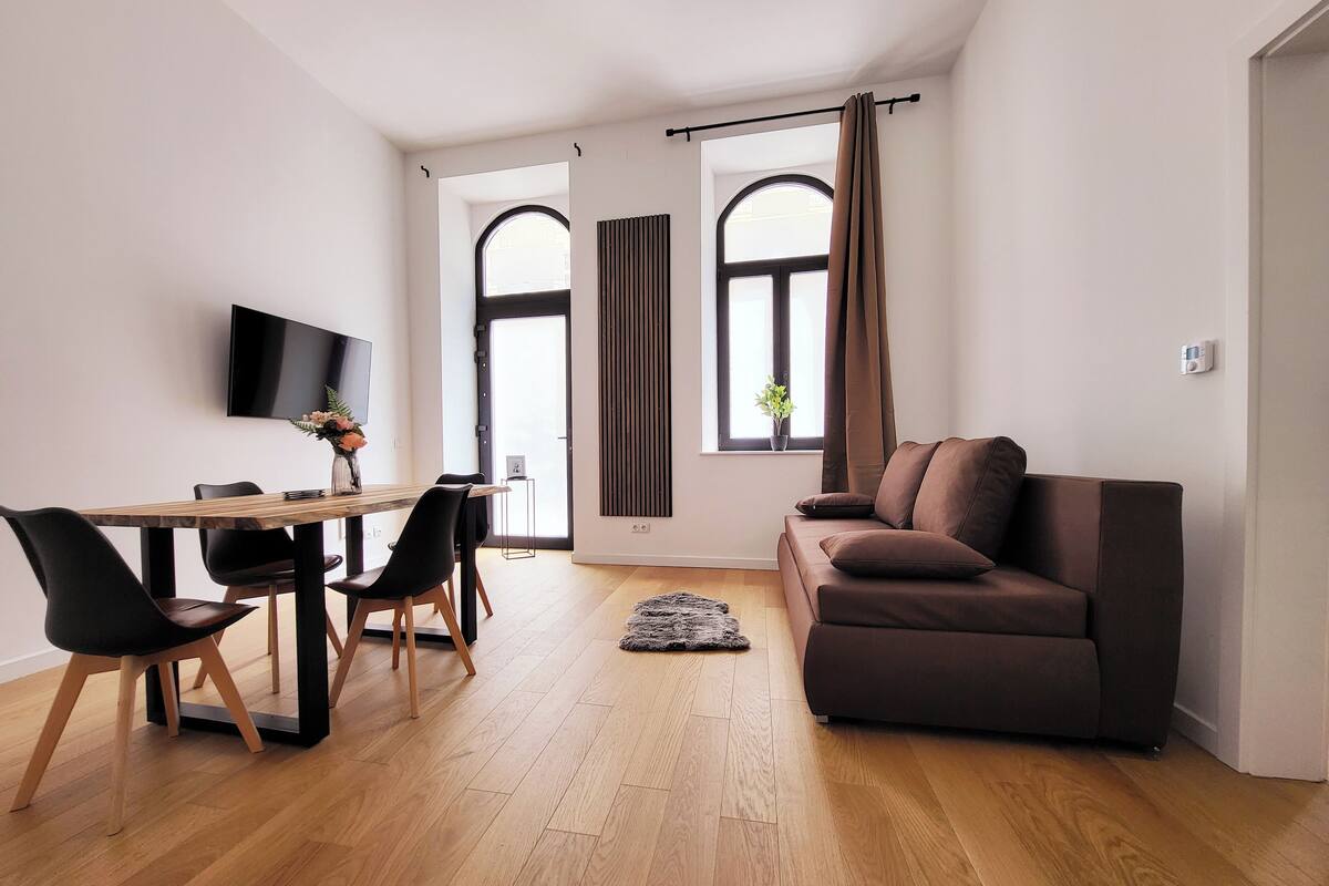 A modern living room with a wooden dining table, black chairs, a brown sofa, and a flat-screen TV mounted on the wall.