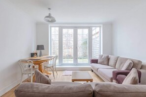 A bright living room with a sofa, a coffee table, and a desk with a chair. There is a large window with a view of a brick building outside. - Studio W/private Patio Heart of Brixton, London! (London)