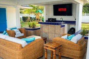An outdoor patio area with wicker furniture, a TV mounted above a tiled bar area, and a children's play structure in the background. - The Heart Villa - Sleeps 16, 5BR Heart-Shaped Pool, Gazebo, Play Park (Canaan, Tobago)