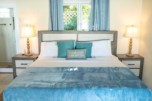 A bed with a blue blanket, two nightstands with lamps, a window with curtains, and a wall-mounted light switch.