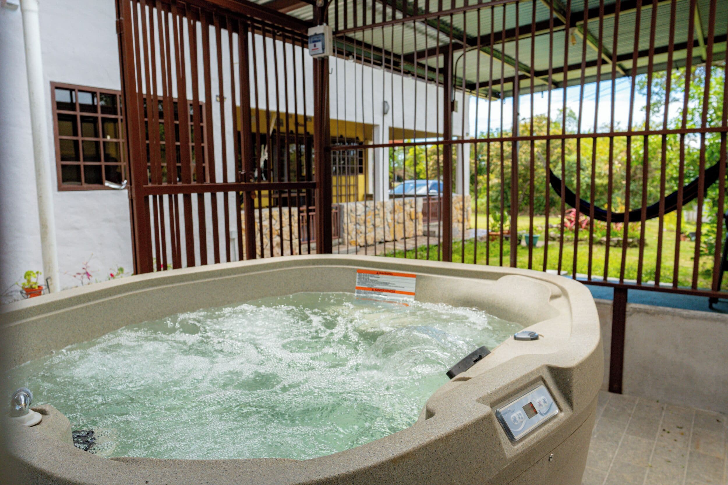 A hot tub with a control panel, surrounded by a metal fence and a view of a house and greenery.