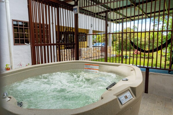A hot tub with a control panel, surrounded by a metal fence and a view of a house and greenery.