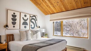 A bedroom with a wooden ceiling, a large bed with white bedding, a nightstand, a bedside table with a lamp, and a window with a view of trees. - Grand Teton View!!! Jackson Hole's Spring Creek Lookout (Jackson)