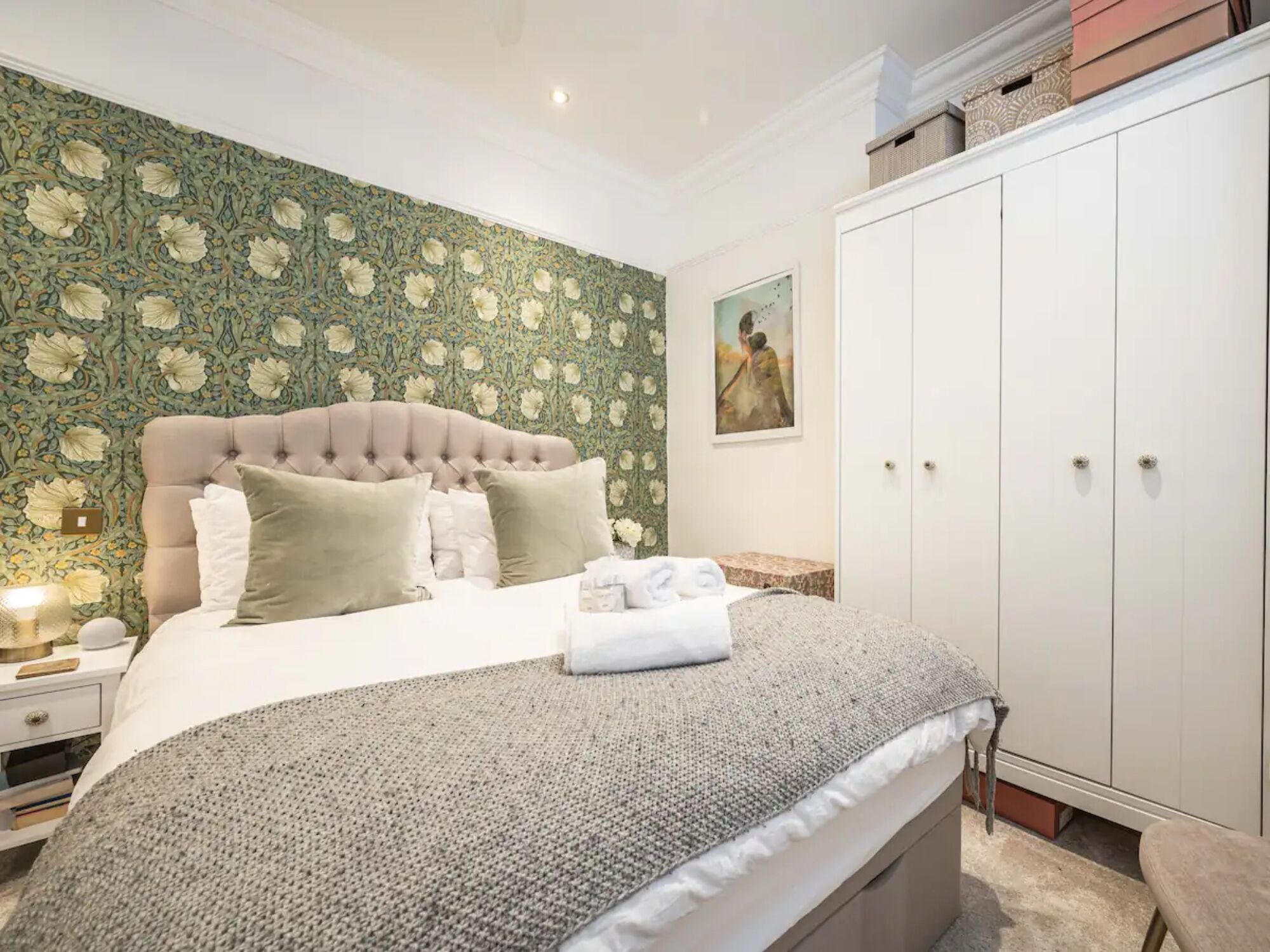 Un dormitorio con cabecera capitonada, cama con manta de punto gris, mesita de noche con lámpara y armario con espejo.