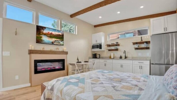 Un dormitorio con una cama grande, televisor montado en la pared, chimenea y una zona de cocina con armarios y electrodomésticos.