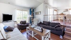 Ein geräumiger Wohnbereich mit einer Couch, zwei Sesseln, einem Couchtisch und einem Fernseher. Es gibt ein Bücherregal, einen Essbereich mit Tisch und Stühlen sowie große Fenster, die für natürliches Licht sorgen.